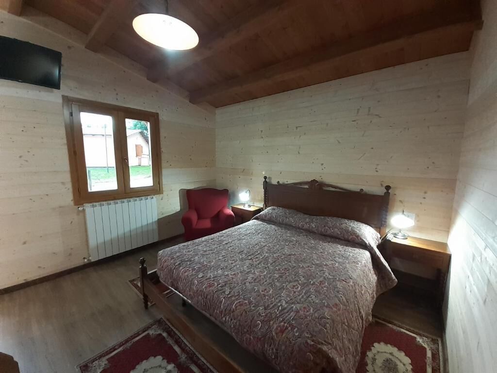 Une chambre avec un plafond en bois, un lit recouvert d’une couvre-lit à motifs, une chaise rouge, une table de chevet avec une lampe et une fenêtre donnant sur une maison.