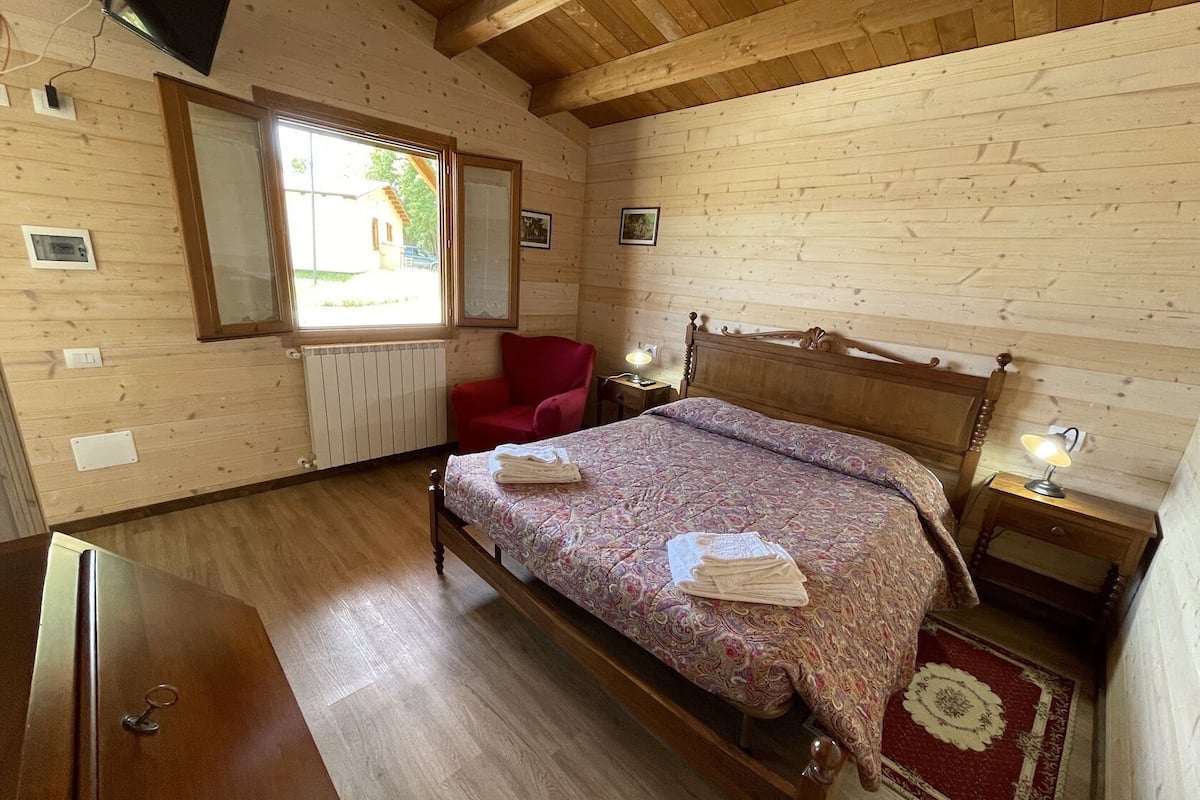A wooden-paneled bedroom with a bed, a red chair, a bedside table with a lamp, and a window with a view of the outside.