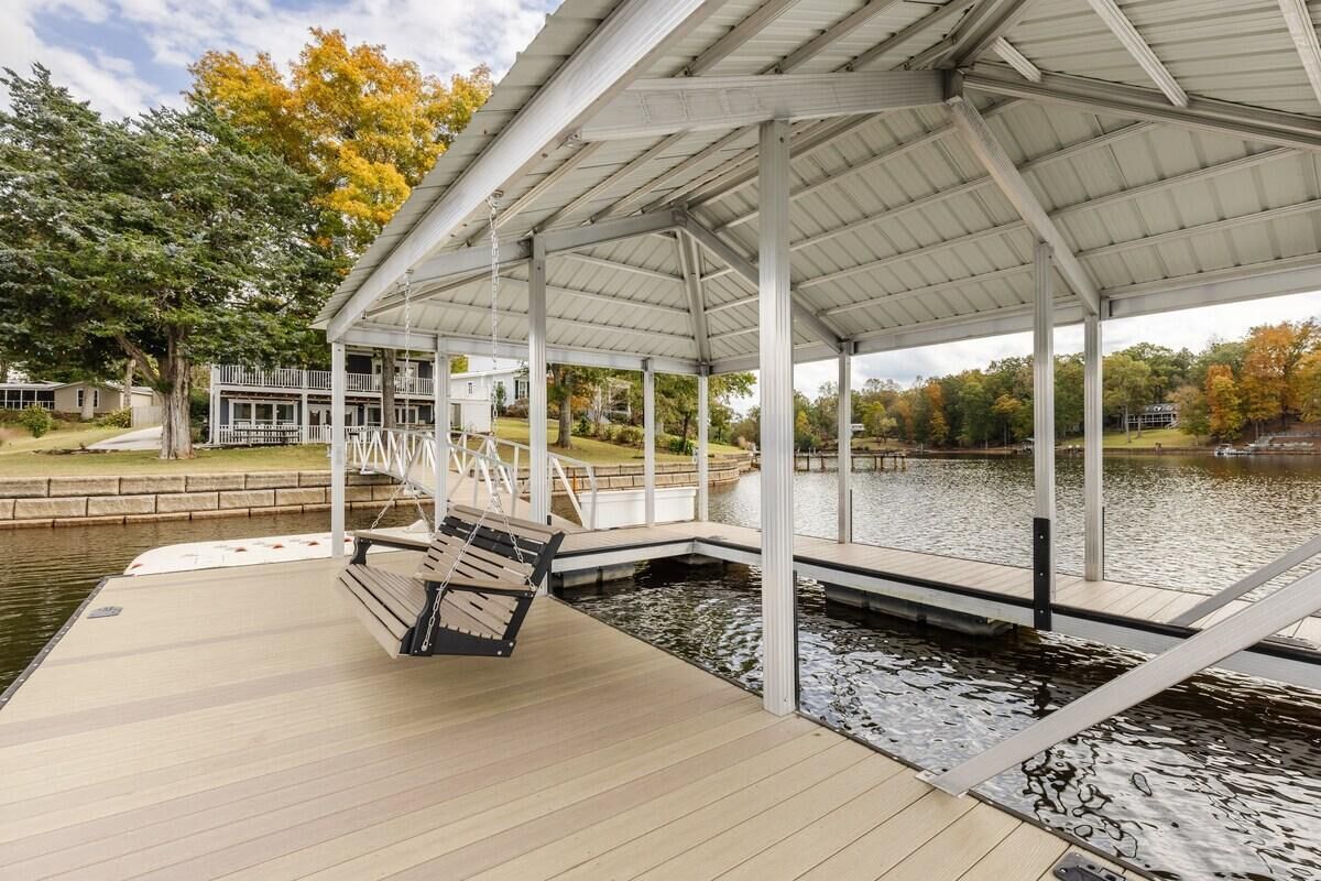 A covered dock with a swing chair, a view of a lake, and a building in the background.