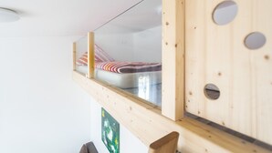 Un lit superposé avec une structure en bois et un panneau en verre, un mur en bois avec des découpes circulaires, et un tableau encadré dans le coin inf érieur gauche.