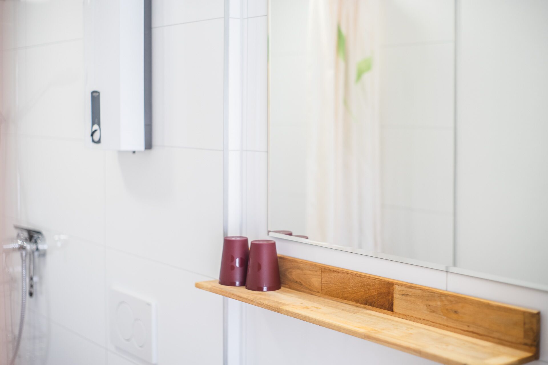 A bathroom with a wooden shelf holding three red cups, a mirror, and a shower area.