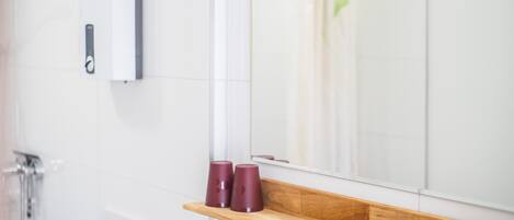 A bathroom with a wooden shelf holding three red cups, a mirror, and a shower area.