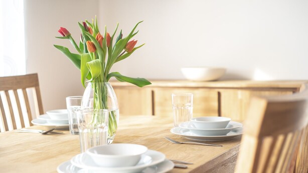 Une table à manger avec de l’assiette, des bols et des couverts blancs, un vase rempli de fleurs et un verre d’eau transparent.