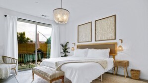 A bedroom with a large bed, a bedside table with a lamp, a wicker chair, and a view of a balcony. - Sunset Oasis: GameRoom, Heated Pool, Hot Tub Gate Community (Indio)