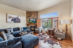 A modern living room with a fireplace, a large sofa, a television, and a view of the outdoors through large windows.