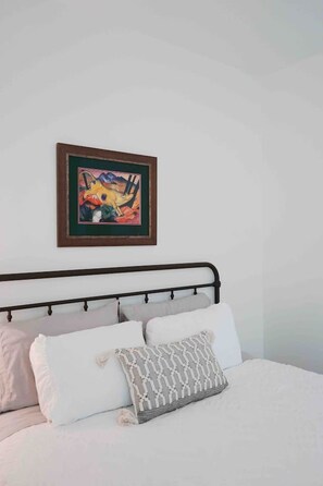 Een bed met wit beddengoed en meerdere kussens, een lijst met abstract schilderij erboven, en een effen witte muur.