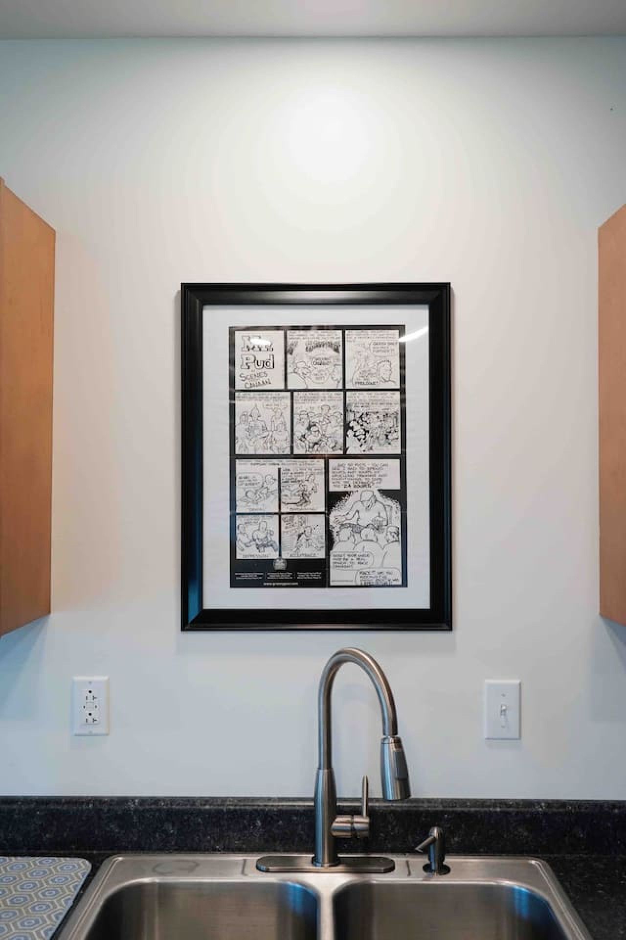 A kitchen with a stainless steel sink, a framed comic strip on the wall, and a wooden cabinet to the left.