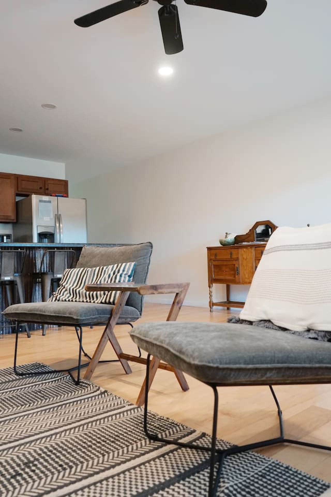 A modern living room with a sofa, armchair, and a dining area with a refrigerator and cabinets.