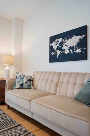 Een woonkamer met een beige bank, een wereldkaart kunstwerk, een kussen met een patroon, een lamp en een vloerkleed met een herringbone-motief.