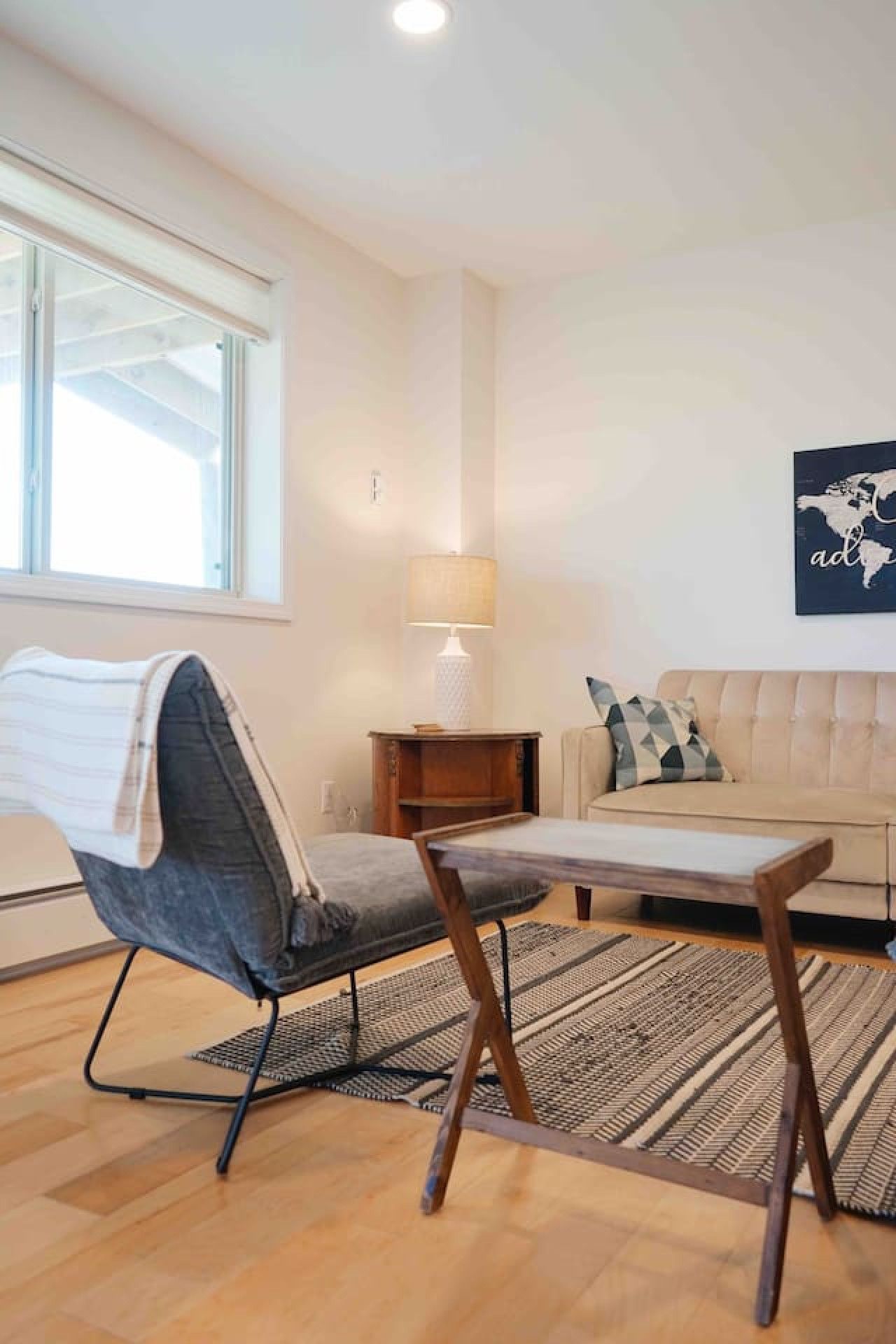 Una habitación con piso de madera, un sofá, una silla, una mesita, una lámpara y un cuadro con un mapa del mundo en la pared.