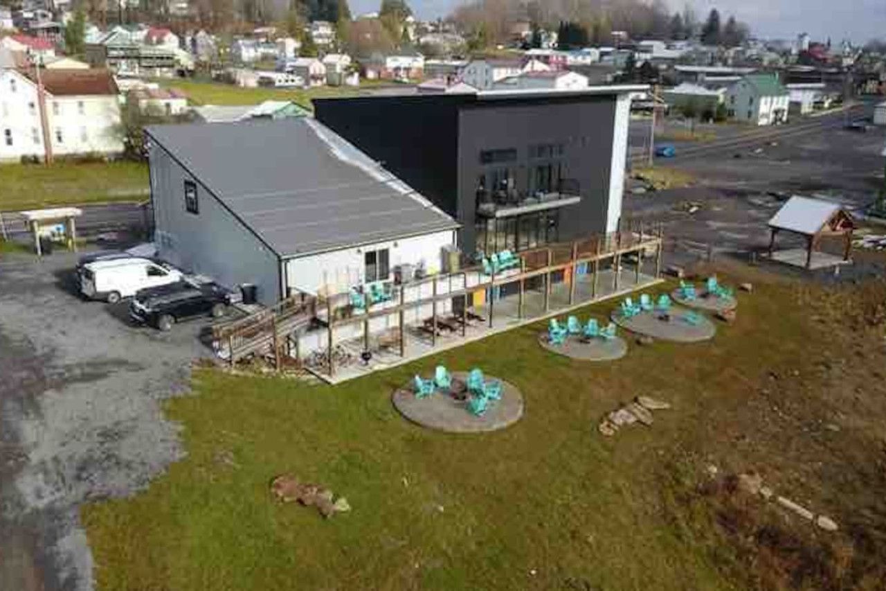 A building with a flat roof, a covered outdoor seating area with tables and chairs, and a parking lot with several vehicles.