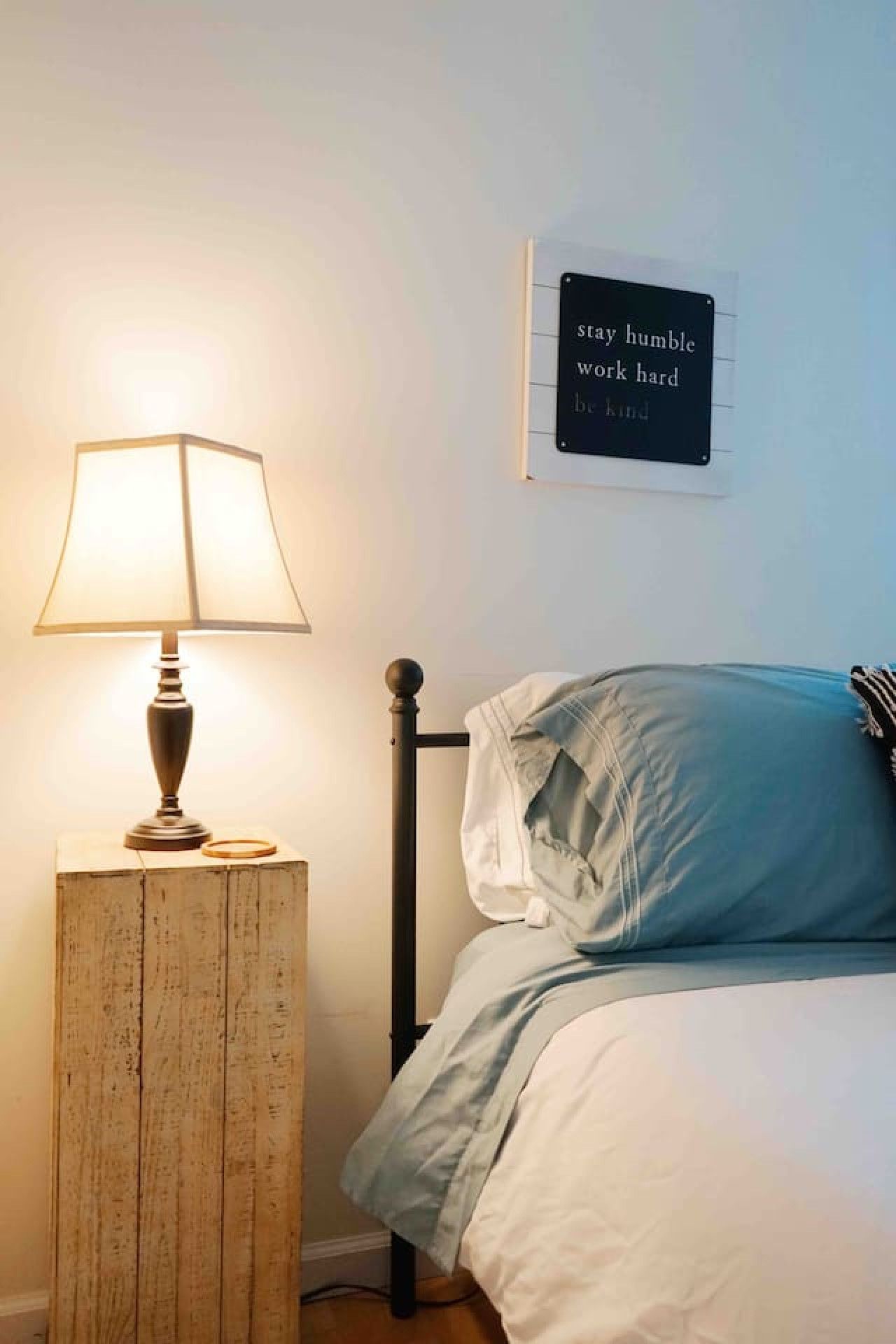 Un dormitorio con una lámpara de noche, una cama con ropa de cama blanca y una frase motivacional en la pared.