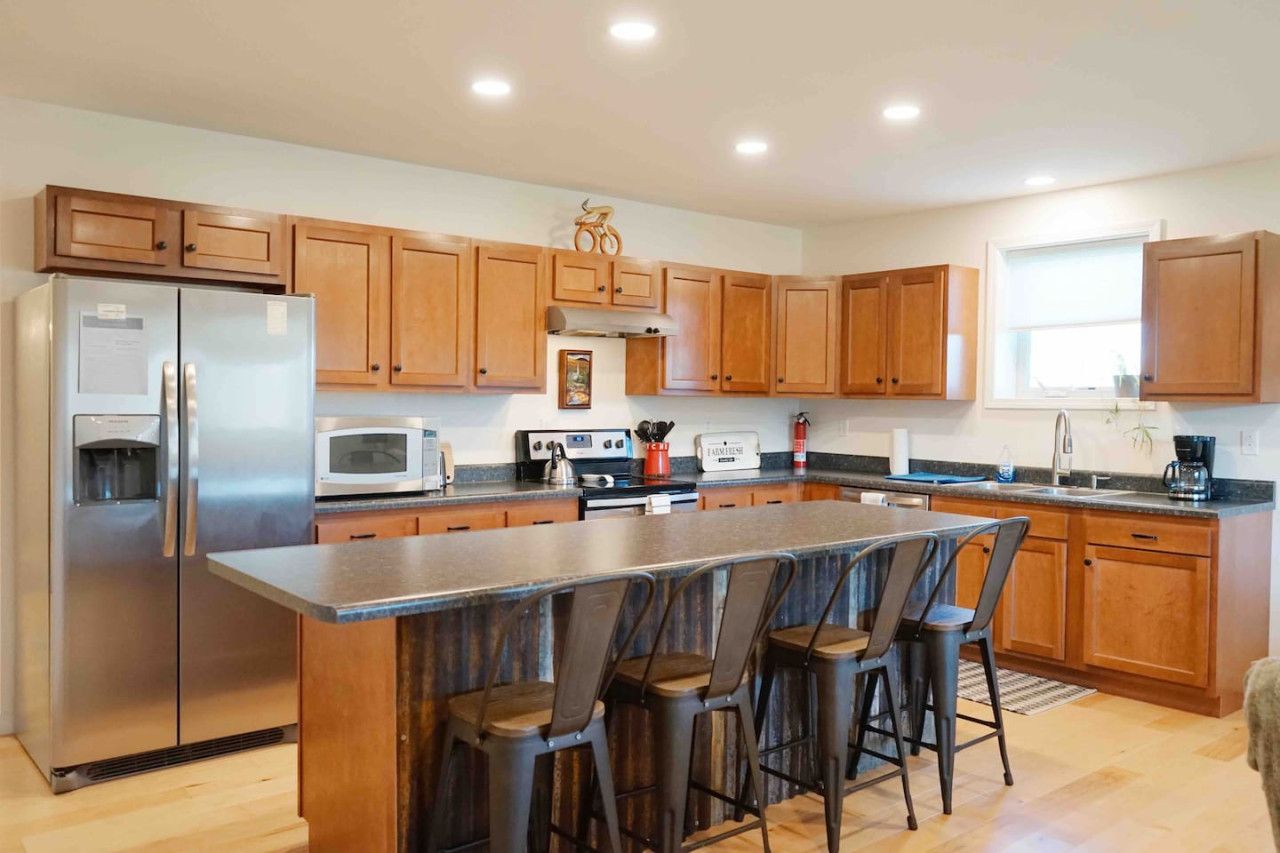 Una cocina moderna con armarios de madera, un refrigerador de acero inoxidable, un microondas, una cafetera y una zona de asientos con taburetes de bar.