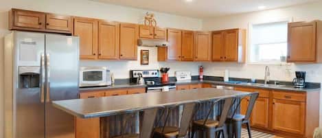 Una cocina moderna con armarios de madera, un refrigerador de acero inoxidable, un microondas, una cafetera y una zona de asientos con taburetes de bar.