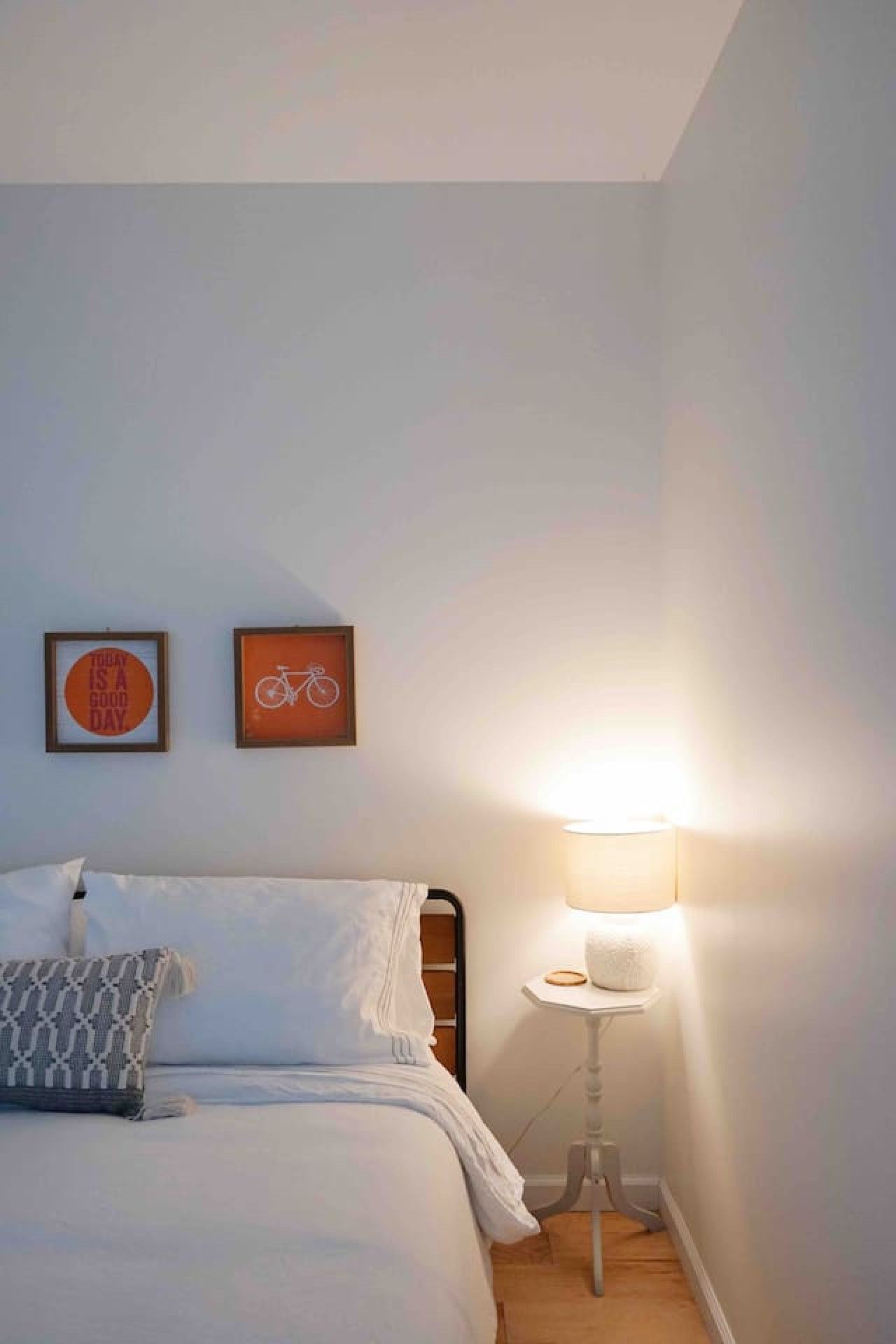 Ein Schlafzimmer mit einem Bett, Kissen, einem Nachttisch mit Lampe sowie zwei gerahmten Bildern an der Wand.