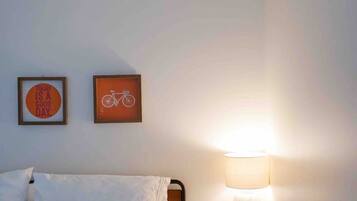 Ein Schlafzimmer mit einem Bett, Kissen, einem Nachttisch mit Lampe sowie zwei gerahmten Bildern an der Wand.