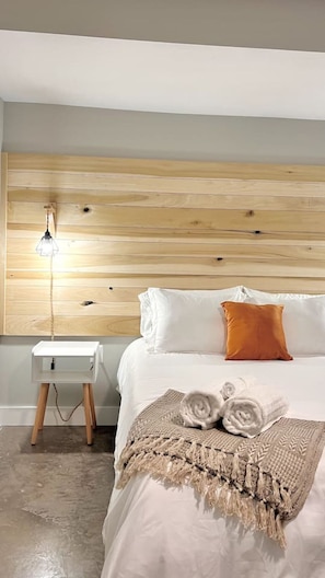 Ein Schlafzimmer mit Holzkopfstütze, einem Nachttisch mit Lampe, weißer Bettwäsche und einem aufgerollten Handtuch auf dem Bett.
