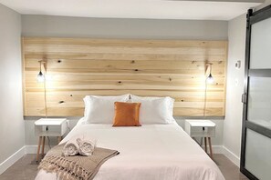 A hotel room with a wooden headboard, two bedside tables, a bed with white bedding and an orange pillow, and a small white nightstand.