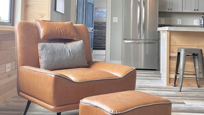 Un salon moderne avec un canapé et un pouf en cuir, une cuisine équipée d’appareils électroménagers en acier inoxydable et un plafond en bois.