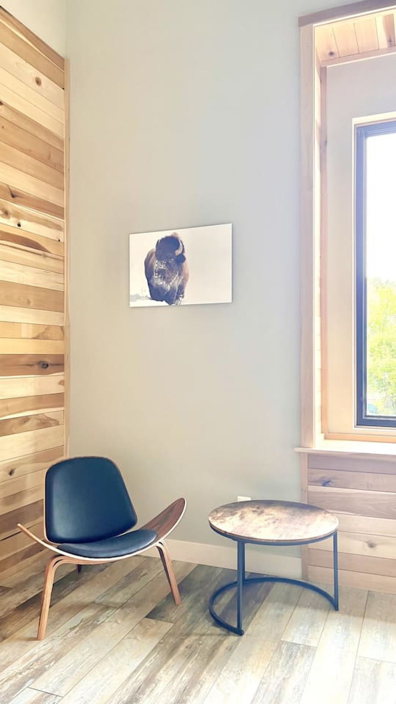 A room with a wooden wall, a blue chair, a small round table, and a window with a view.