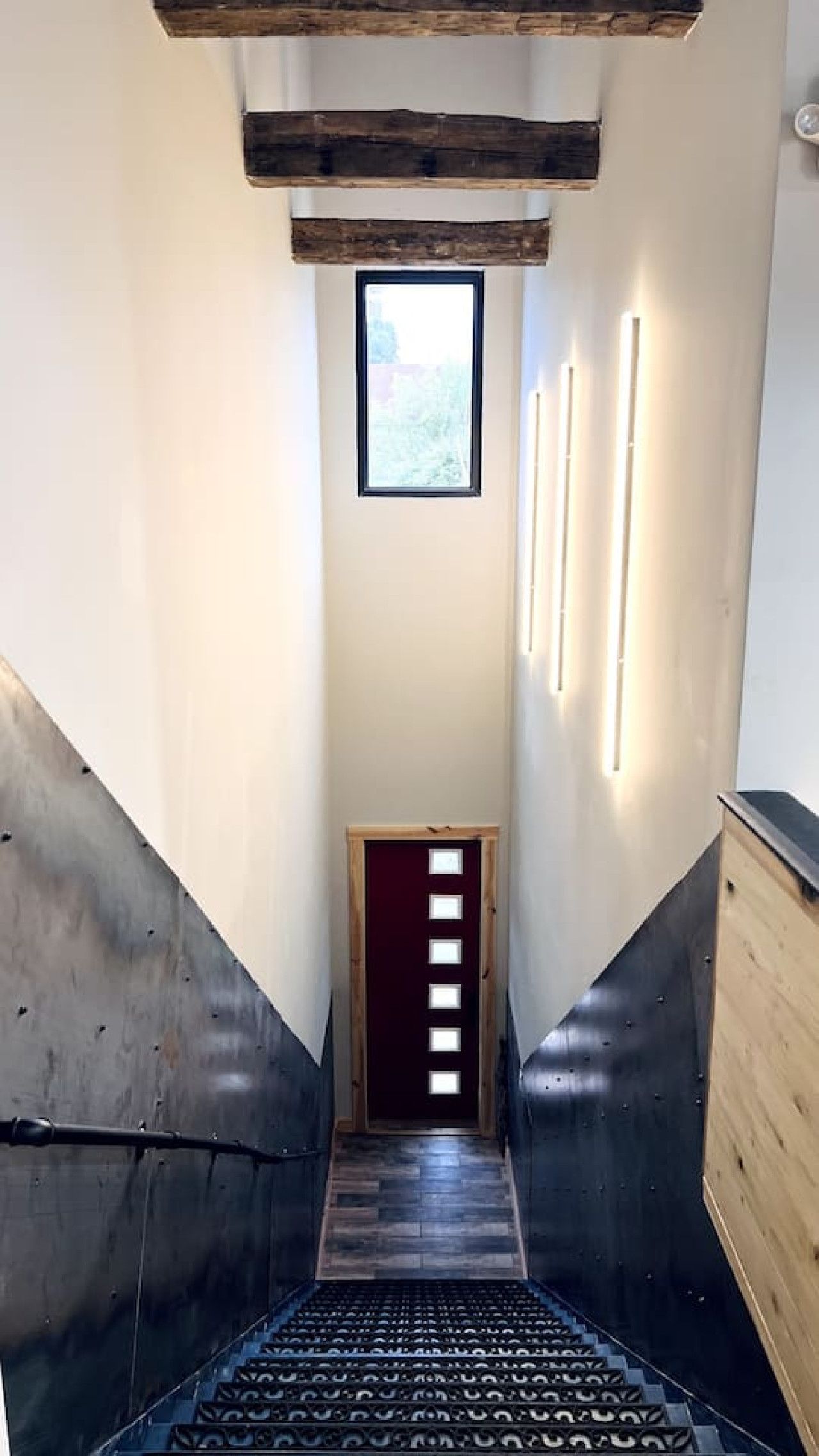 Een smalle trap met een houten leuning, een donker vloerkleed en een deur met een verticale lichtbalk.