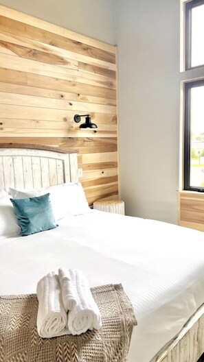 Una cama bien tendida con sábanas blancas y una almohada azul, un cabecero de madera y una ventana con vistas.
