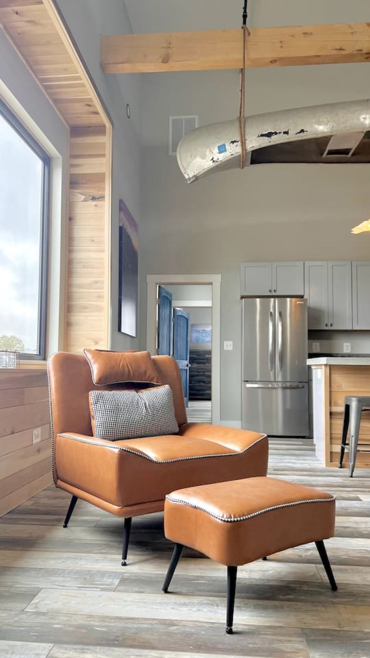 Een moderne woonkamer met een leren bank en voetenbankje, een houten plafond en uitzicht op de keuken.