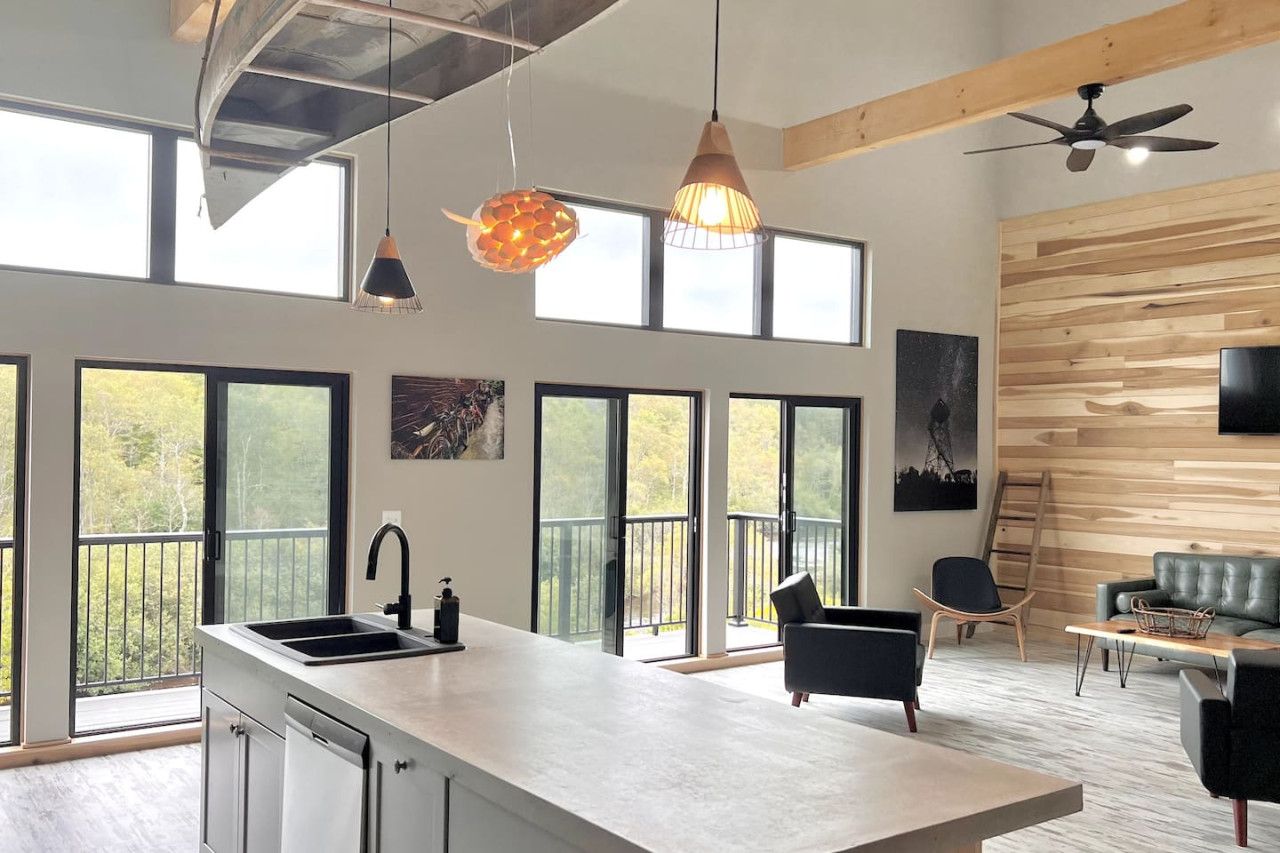 A modern kitchen with a large island, a dining area with a sofa and chairs, and a balcony with a view of trees.