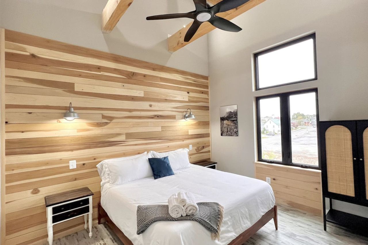 Ein Schlafzimmer mit einer Wand aus Holzlatten, einem Bett mit weißer Bettwäsche, einem Deckenventilator, einem Fenster mit Blick und einem Nachttisch.