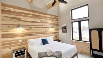 Ein Schlafzimmer mit einer Wand aus Holzlatten, einem Bett mit weißer Bettwäsche, einem Deckenventilator, einem Fenster mit Blick und einem Nachttisch.