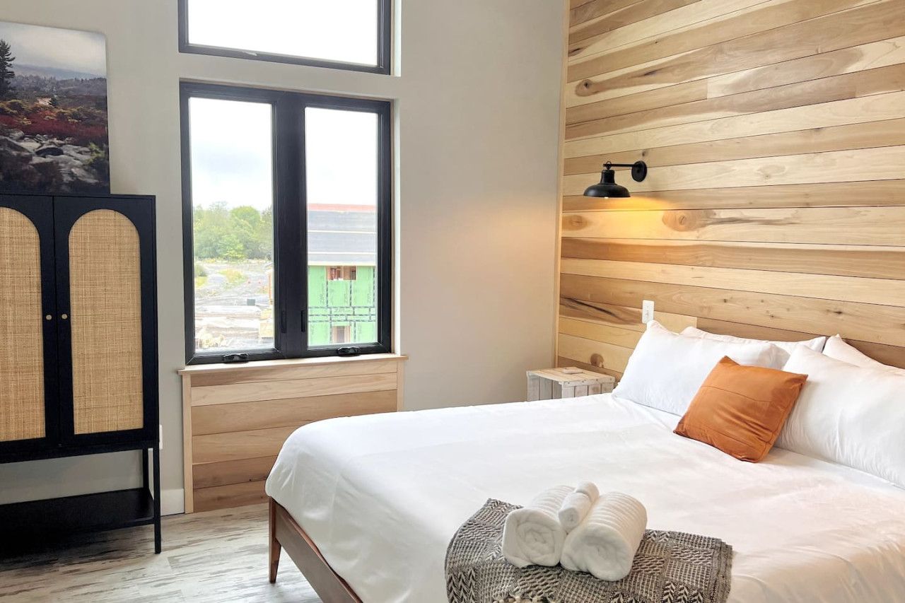 A hotel room with a large bed, a window with a view, a wooden wall, and a nightstand with a lamp.