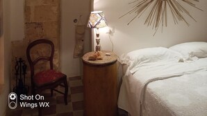 A room with a stone wall, a round wooden table with a lamp, a chair, and a bed with white bedding. - loft Salento Style (Alessano)
