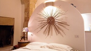 A bedroom with a large bed, a wall-mounted decorative light, a small table with a lamp, and a stone wall. - loft Salento Style (Alessano)