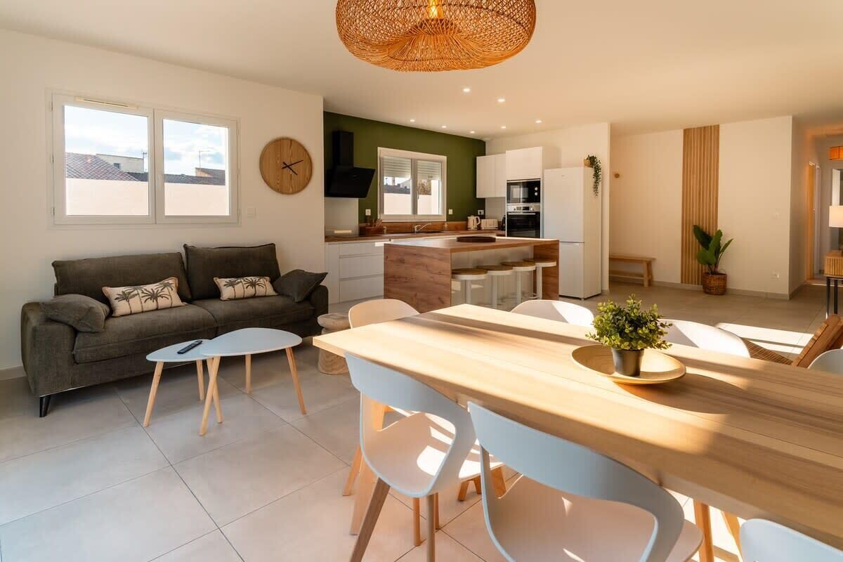 Een moderne woonruimte met een bank, eettafel en stoelen. Er is een keuken met een kookplaat, spoelbak en kastjes. Op de muur hangt een klok.