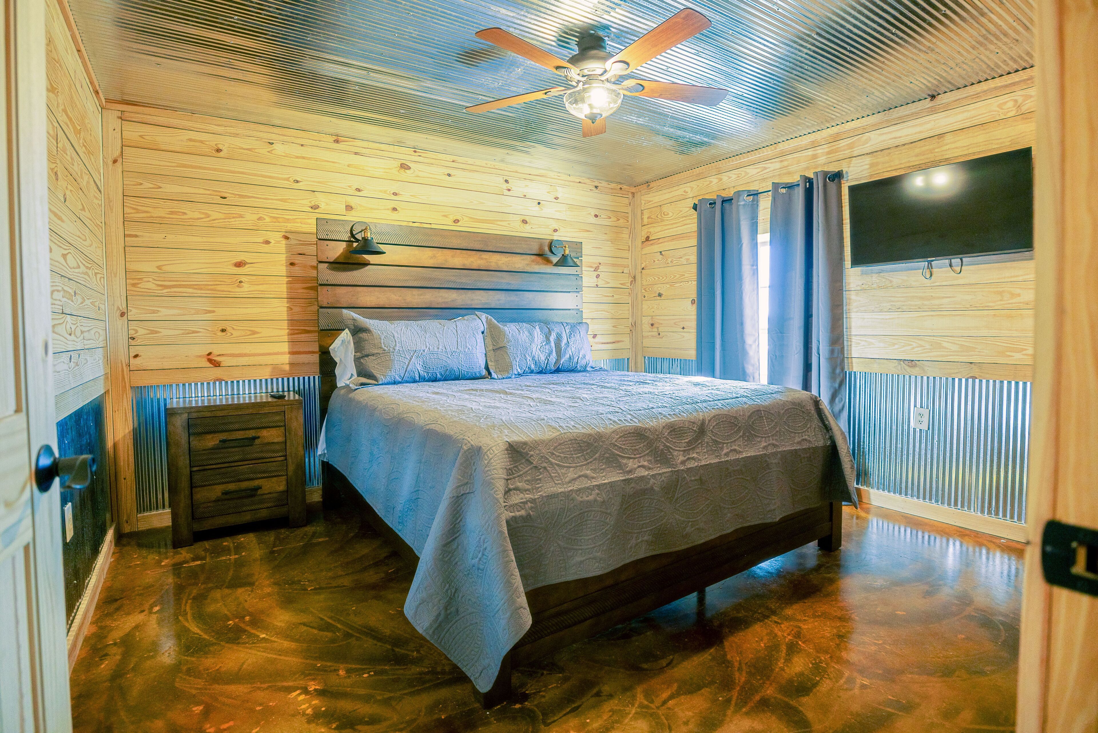 A wooden-paneled bedroom with a bed, a nightstand, a ceiling fan, and a window with curtains.