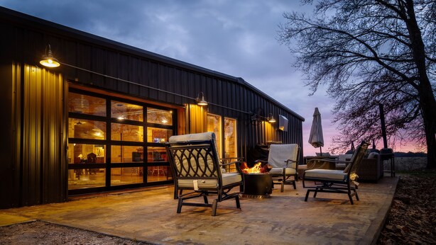 A building with a large garage door, outdoor seating area with chairs and a fire pit, and a tree in the background.