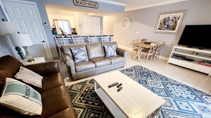Una sala de estar con un sofá de cuero marrón, una mesa de centro blanca y un tapete estampado. Hay un comedor con una mesa y sillas, y un televisor sobre un mueble.