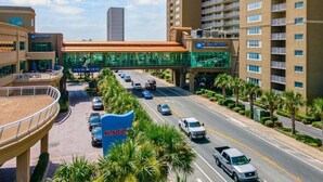 Un hotel de varios pisos con fachada de vidrio, una galería techada y un estacionamiento con automóviles.