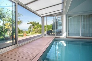 A pool area with a wooden deck, large sliding glass doors, and a view of the sea.