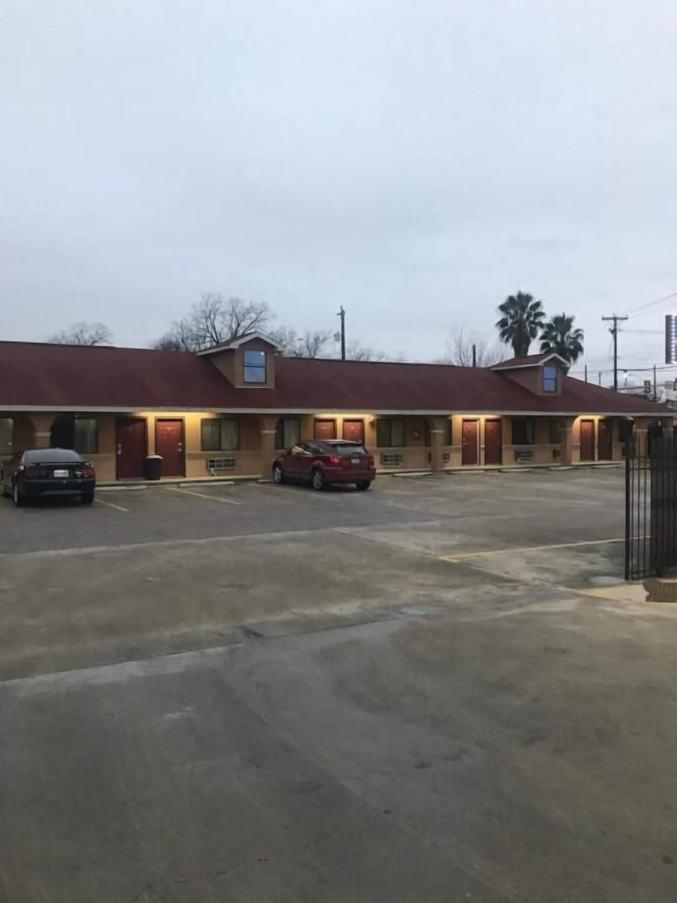 A motel with a red roof and multiple doors, a parking lot with two cars, and a palm tree in the background.