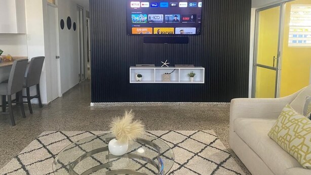Een woonkamer met een flatscreen televisie aan een donkere muur, een glazen salontafel met een decoratieve vaas, en een beige bank met gekleurde kussens.
