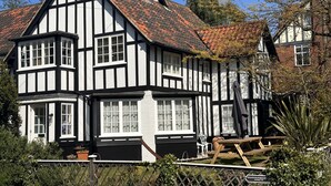 Ett traditionellt hus med korsvirkesfasad, många fönster, ett tak täckt med tegelpannor och en inhägnad trädgård med picknickbord.