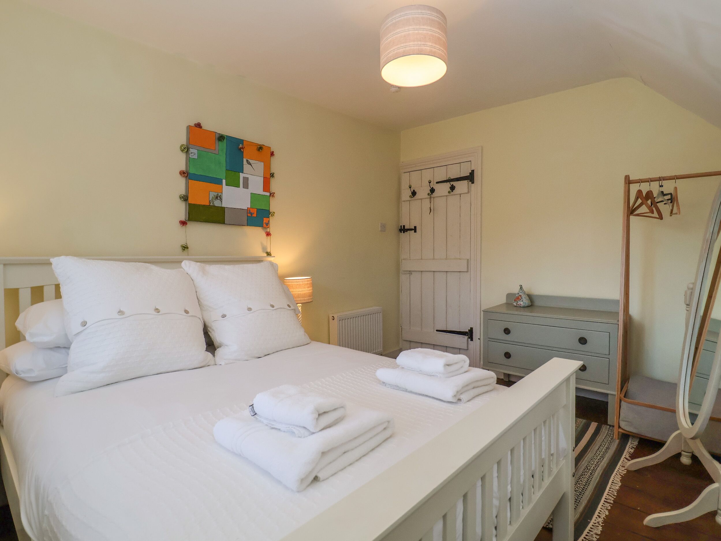 A neatly made bed with white linens and pillows, a colorful artwork on the wall, a white door with a handle, and a wooden chair with a hanger.