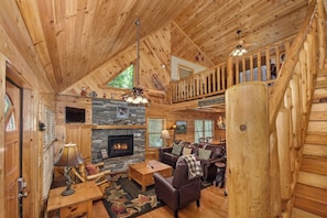 A log cabin interior with a stone fireplace, wooden furniture, and a balcony. - Rustic 3BD+Loft Cabin! HOT TUB + Firepit w/ VIEWS! (Sevierville)