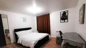 Un dormitorio con cama, escritorio, silla, ventana con cortinas, radiador y un cuadro en la pared.