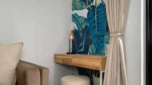 A room with a beige sofa, a wooden desk with a lamp, and a large tropical leaf wallpaper.