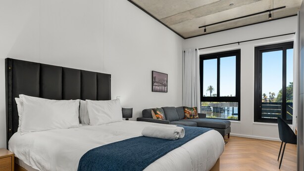 A modern hotel room with a large bed, a sofa, and a view of the outdoors through large windows.