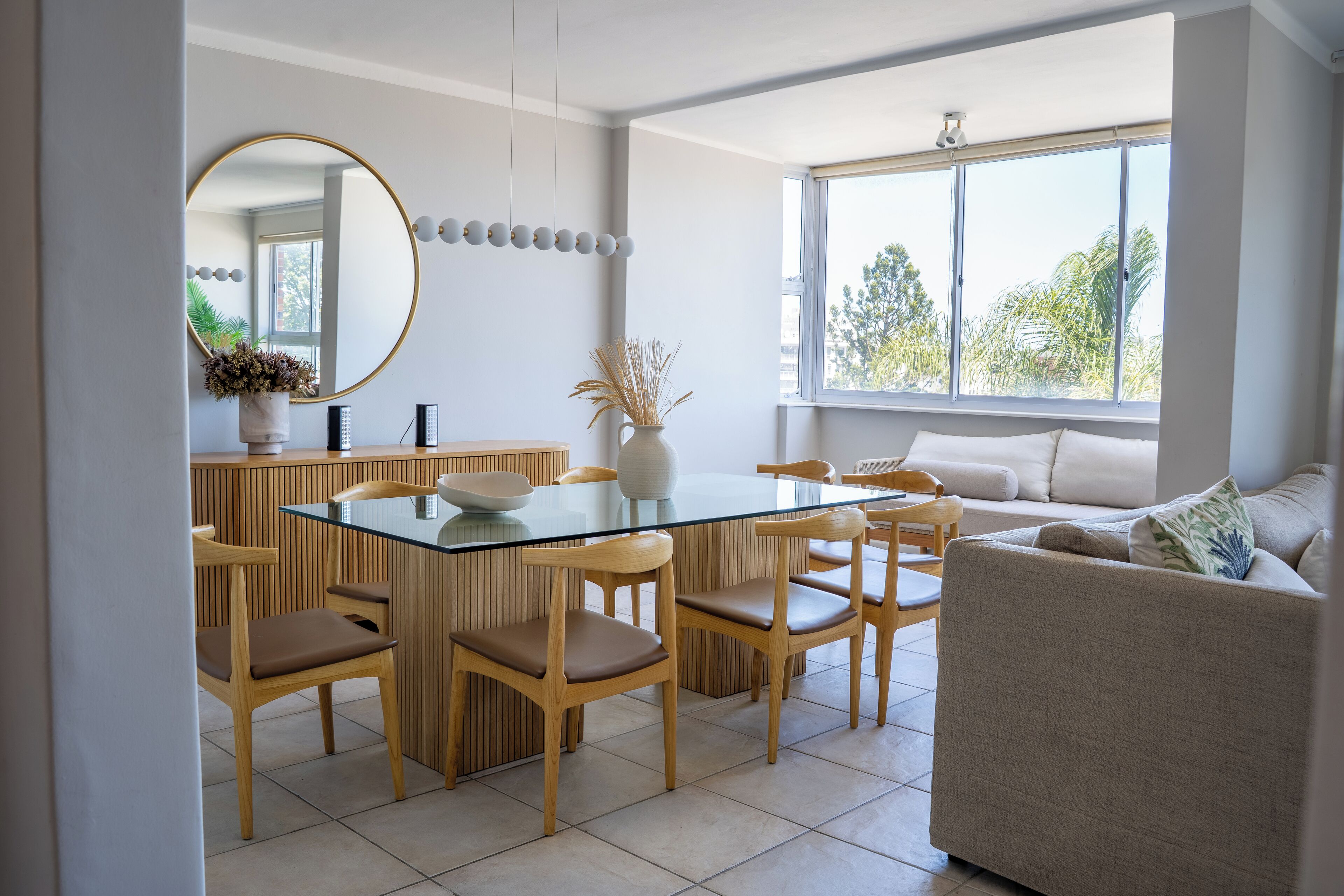 A modern dining area with a glass-top table, wooden chairs, a round mirror, and a large window with a view of trees.