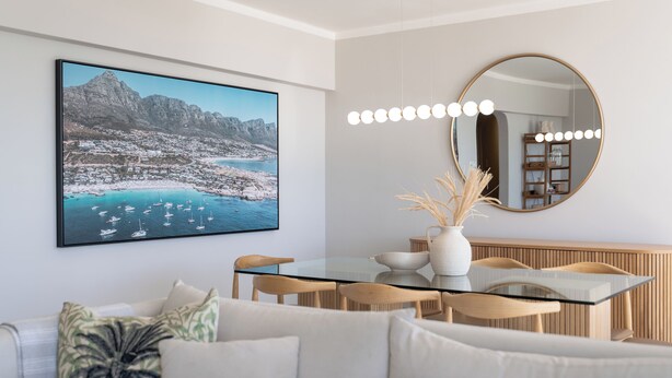 Un espace repas moderne avec une table en verre, des chaises en bois, un vase blanc contenant des plantes séchées et un grand tableau représentant un paysage, encadré et accroché au mur.