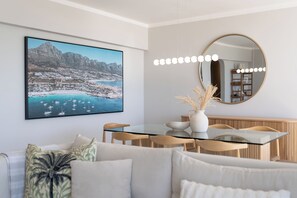 Un espace repas moderne avec une table en verre, des chaises en bois, un vase blanc contenant des plantes séchées et un grand tableau représentant un paysage, encadré et accroché au mur.
